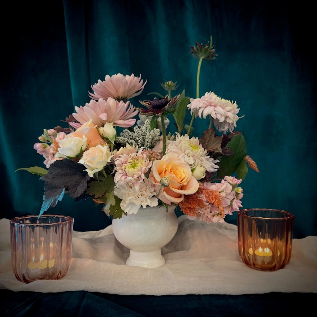 From Campanula Design Studio: A perfect centerpiece for your Thanksgiving table, 'Gratitude' is full of soft blush blossoms complemented with peachier tones and dark accents. Add two of our soft orange votives for a complete centerpiece. 
