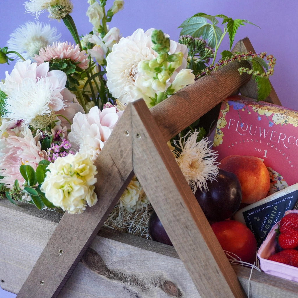Wooden gift basket with flowers, fruits, and a box of chocolates on a wooden surface.