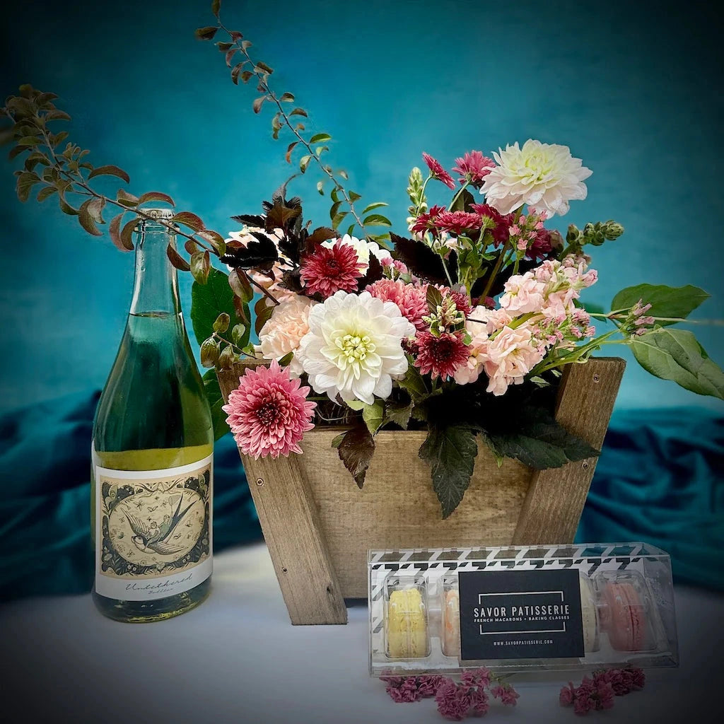 Floral arrangement in a wooden crate with a bottle of wine and a box of macarons on a blue background