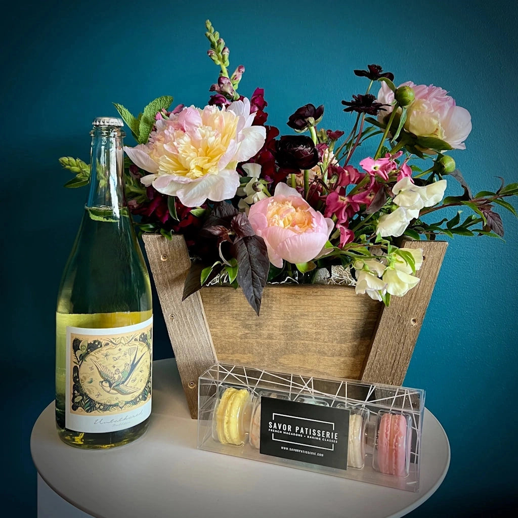 Bottle of wine, floral arrangement, and macaron box on a white surface with a blue background