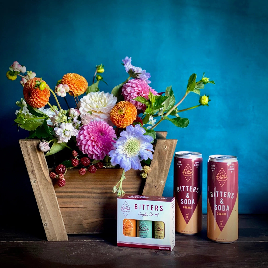 Gift basket featuring a brightly colorful seasonal floral, a sampler set of The Bitter Housewife classic bitters, and two cans of Bitters & Soda - a zero alcohol, refreshing drink. 