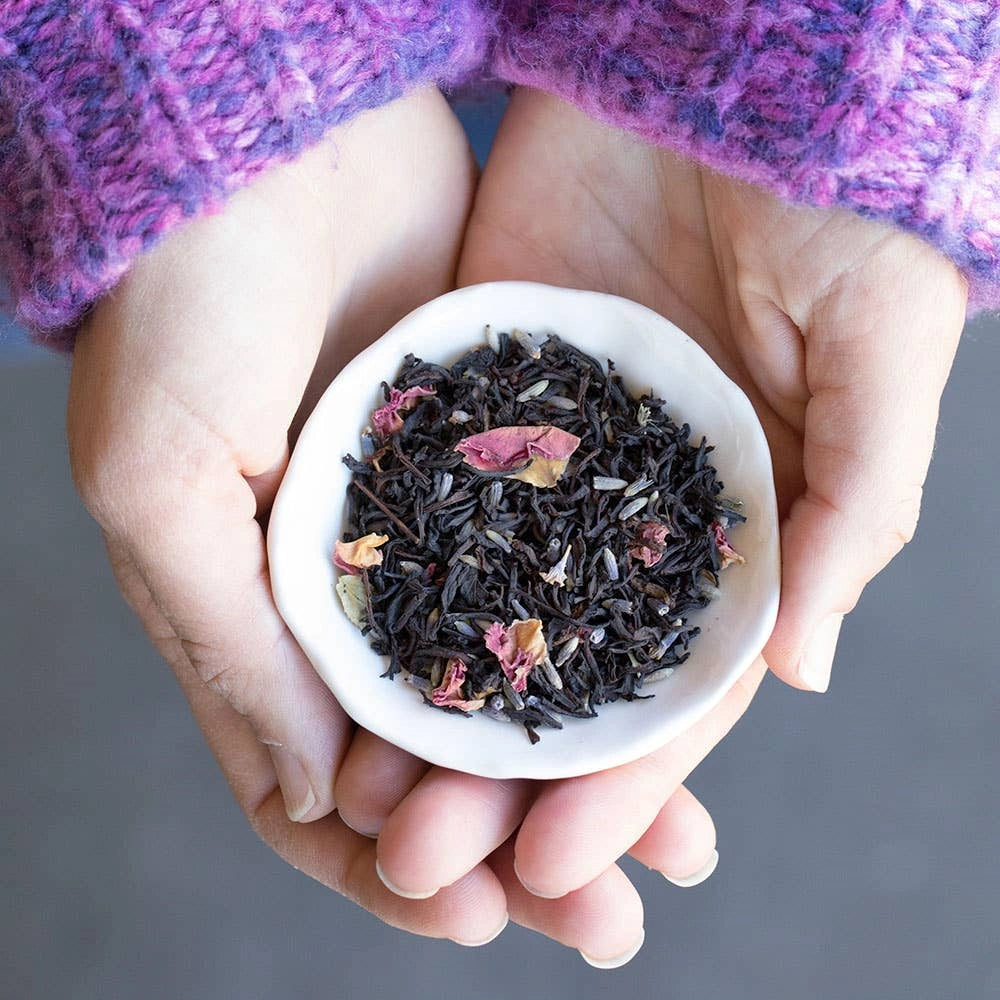 Hands holding a small white dish with lavender black tea leaves and dried flowers against a gray background