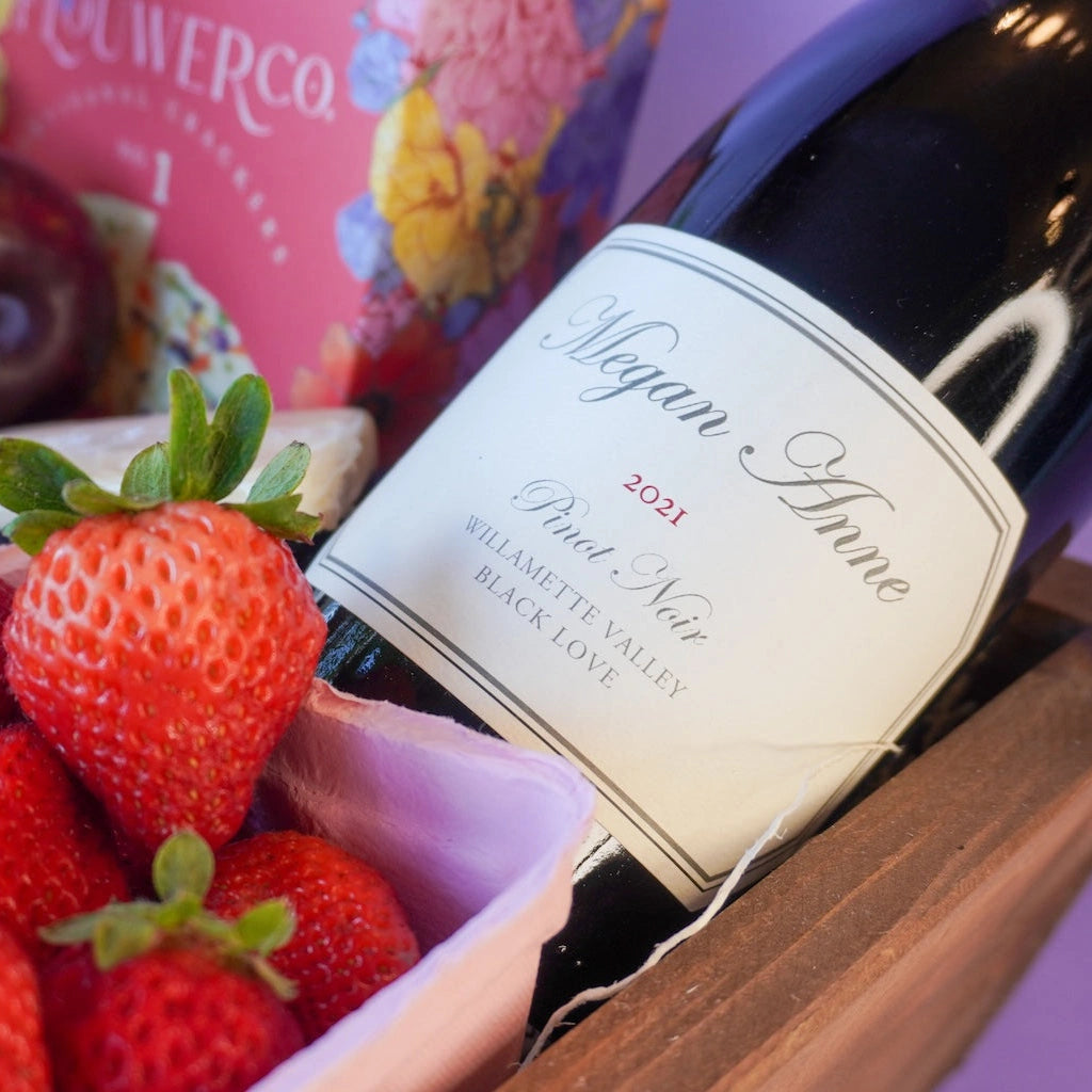 Bottle of Mark Ryan Megan Anne Black Love Pinot Noir wine with a label, surrounded by strawberries and a colorful box in the background.