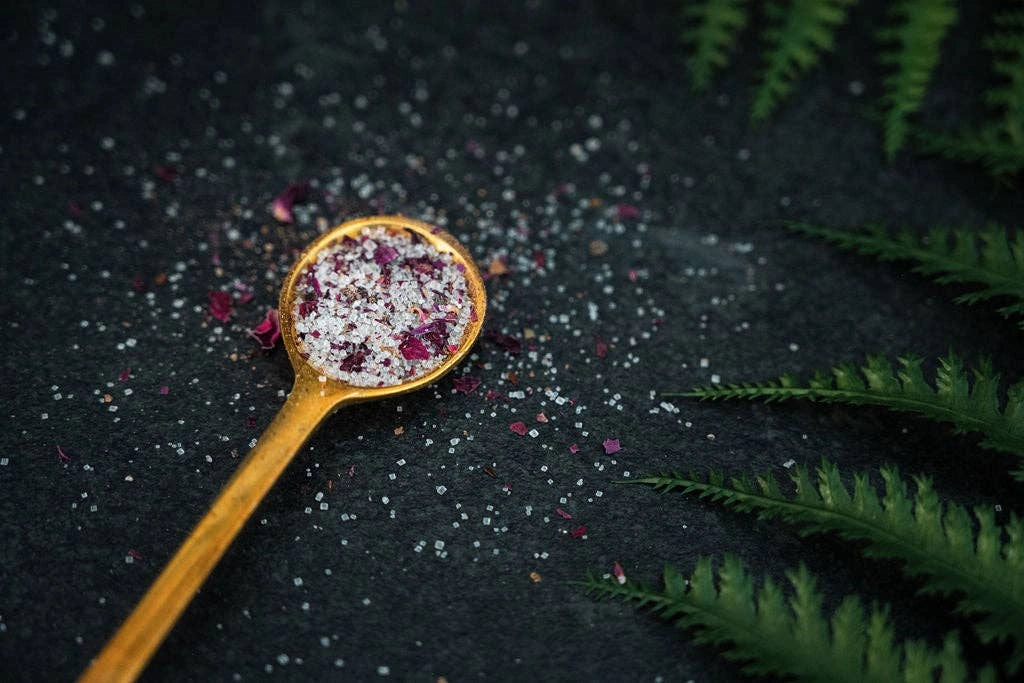 Gold spoon with decorative salt and herbs on a dark surface with fern leaves