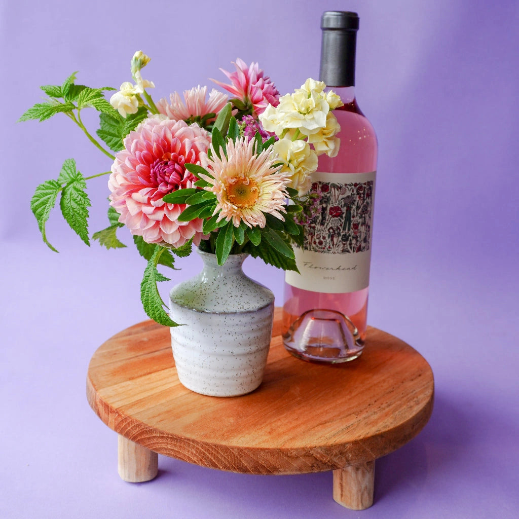 Bouquet of flowers in a vase next to a bottle of rose wine on a wooden stand against a purple background