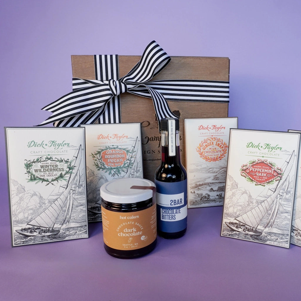 Gift set with chocolate cocktail bitters, hotcakes chocolate syrup, and assorted Dick Taylor craft chocolate bars on a purple background