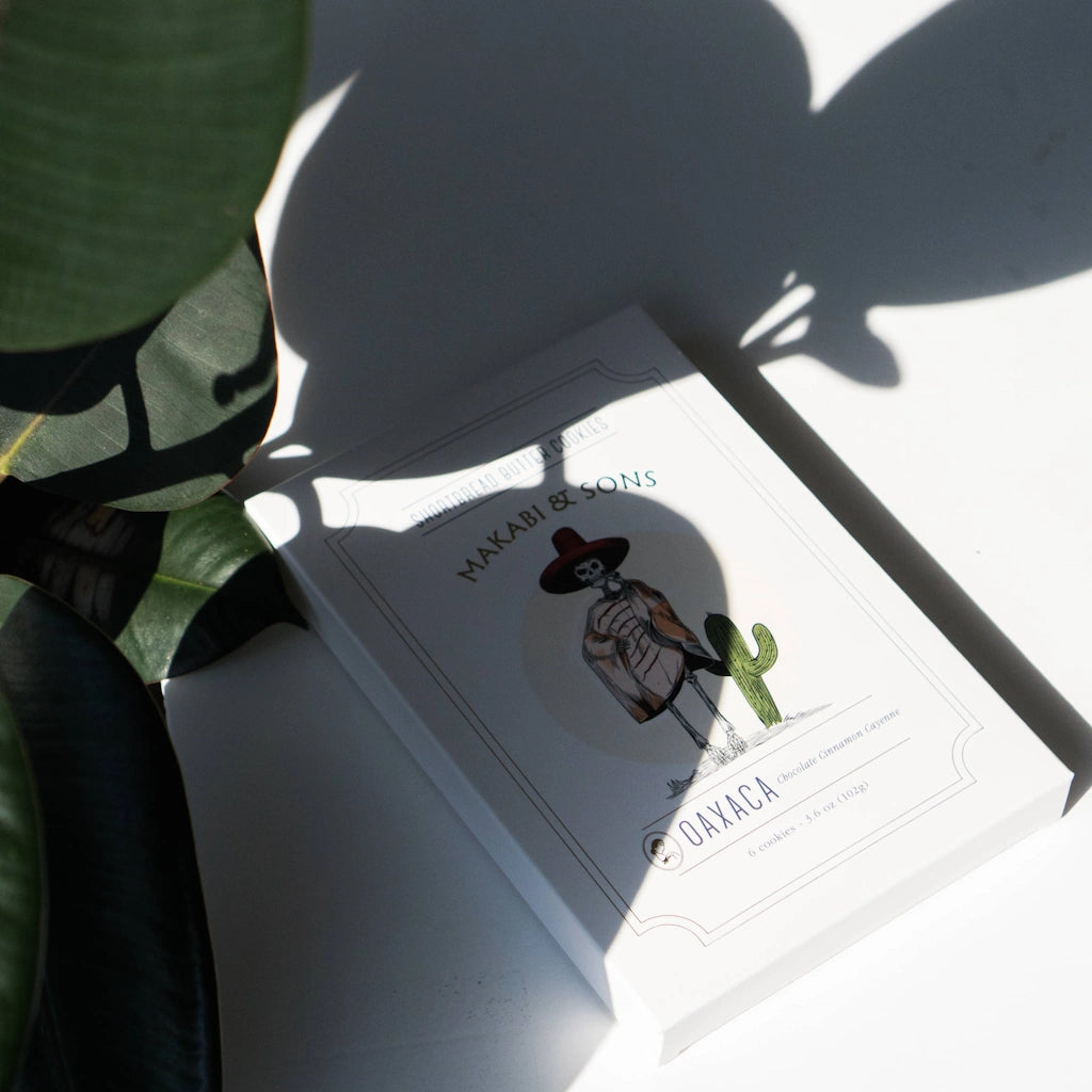 Shortbread cookie box with a skeleton wearing a hat and cactus, placed on a surface with plant shadows.