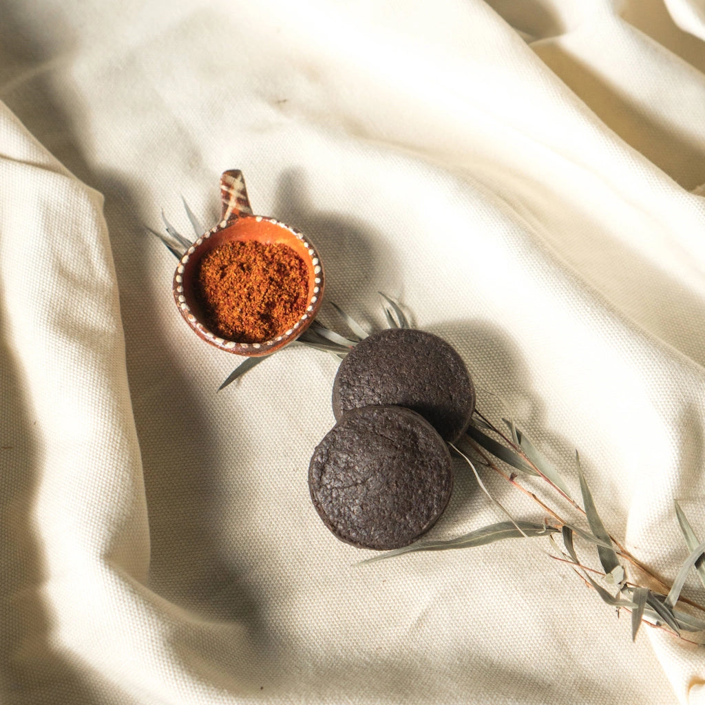 Two dark chocolate shortbread cookies on a light fabric background with a small bowl of spices.