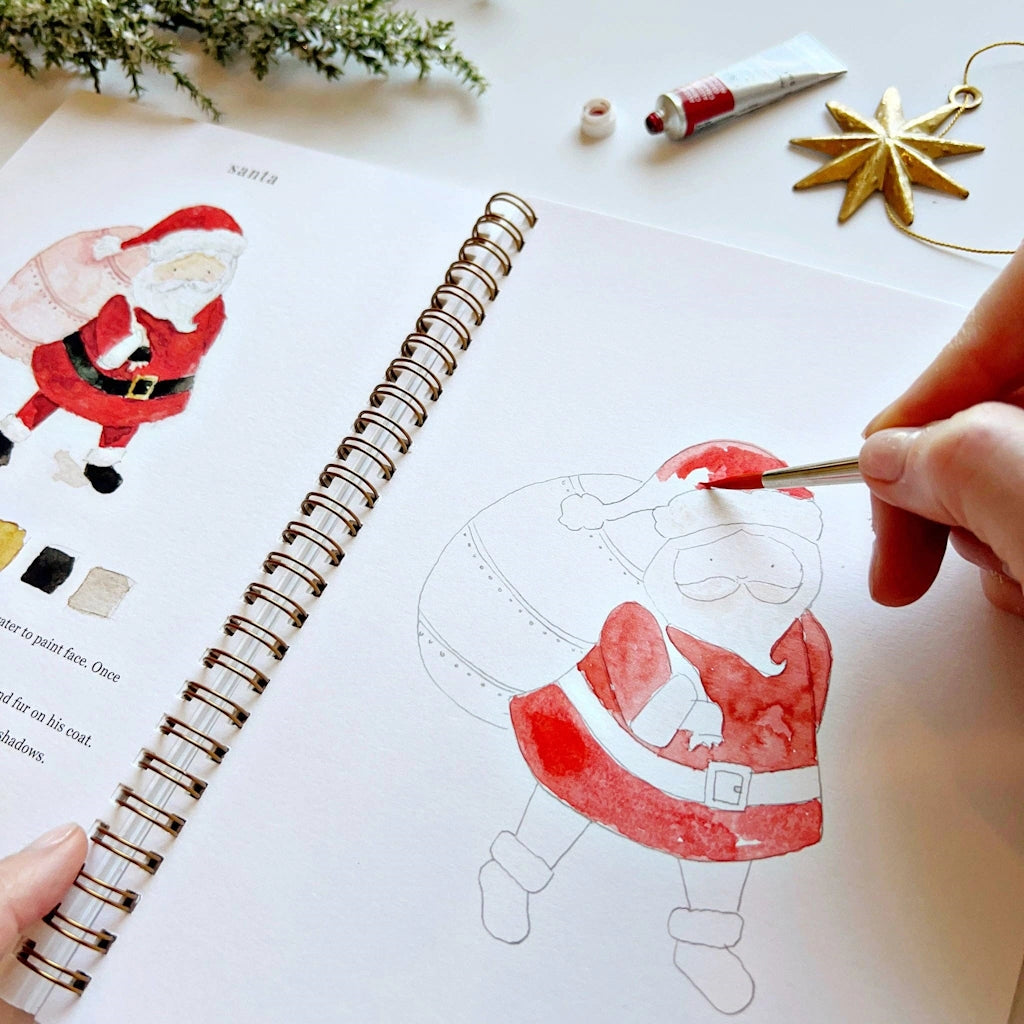 Person coloring a Santa Claus illustration in a spiral-bound book with art supplies around.  Available for sale from Campanula Design Studio, Seattle florist and gift basket company in the Magnolia neighborhood.