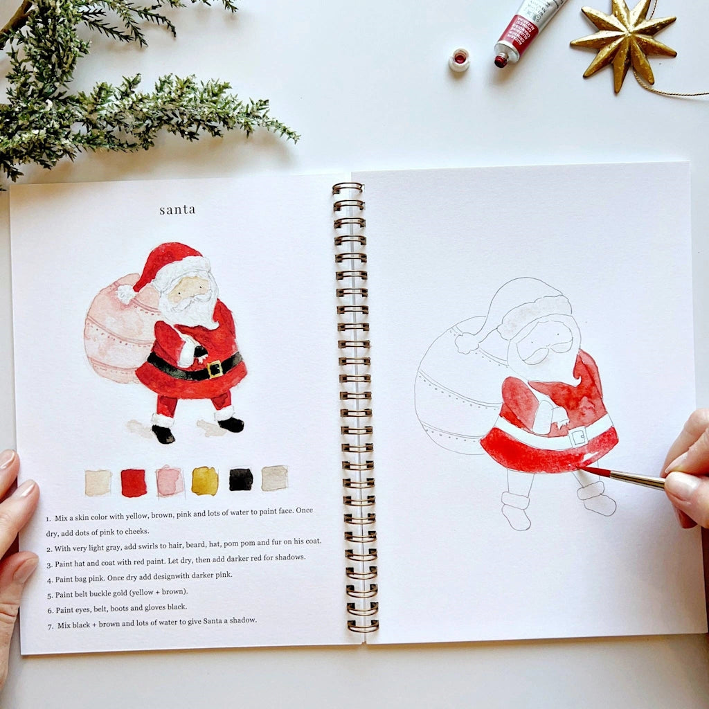 Open book with Santa Claus illustration and painting guide on a white surface with Christmas decorations.  Available for sale from Campanula Design Studio, Seattle florist and gift basket company in the Magnolia neighborhood.
