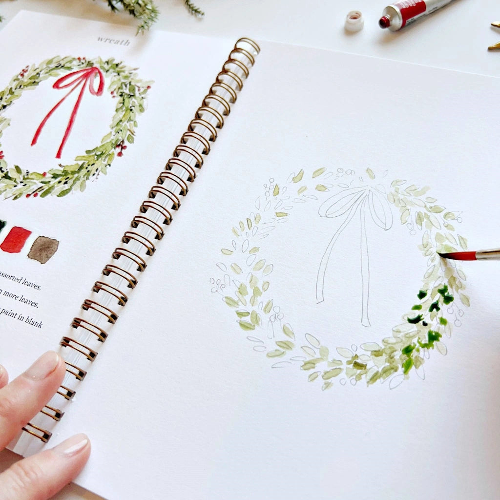 Hand holding a pen over a drawing of a wreath in a spiral-bound notebook.  Available for sale from Campanula Design Studio, Seattle florist and gift basket company in the Magnolia neighborhood.