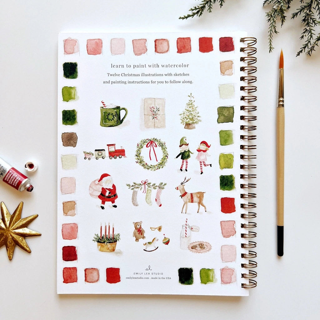 Spiral-bound art book with Christmas illustrations and watercolor swatches on a white surface with a paintbrush and decorative items.  Available for sale from Campanula Design Studio, Seattle florist and gift basket company in the Magnolia neighborhood.
