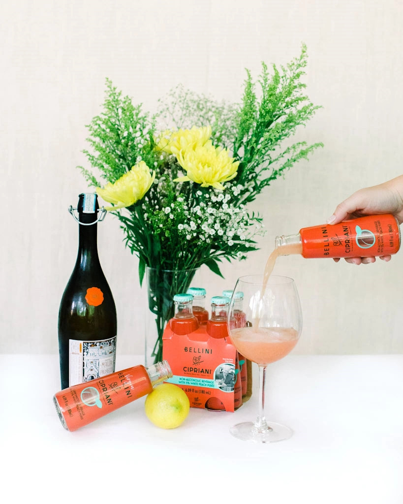 Person pouring a drink from a red bottle into a glass with a bouquet of flowers and bottles in the background.