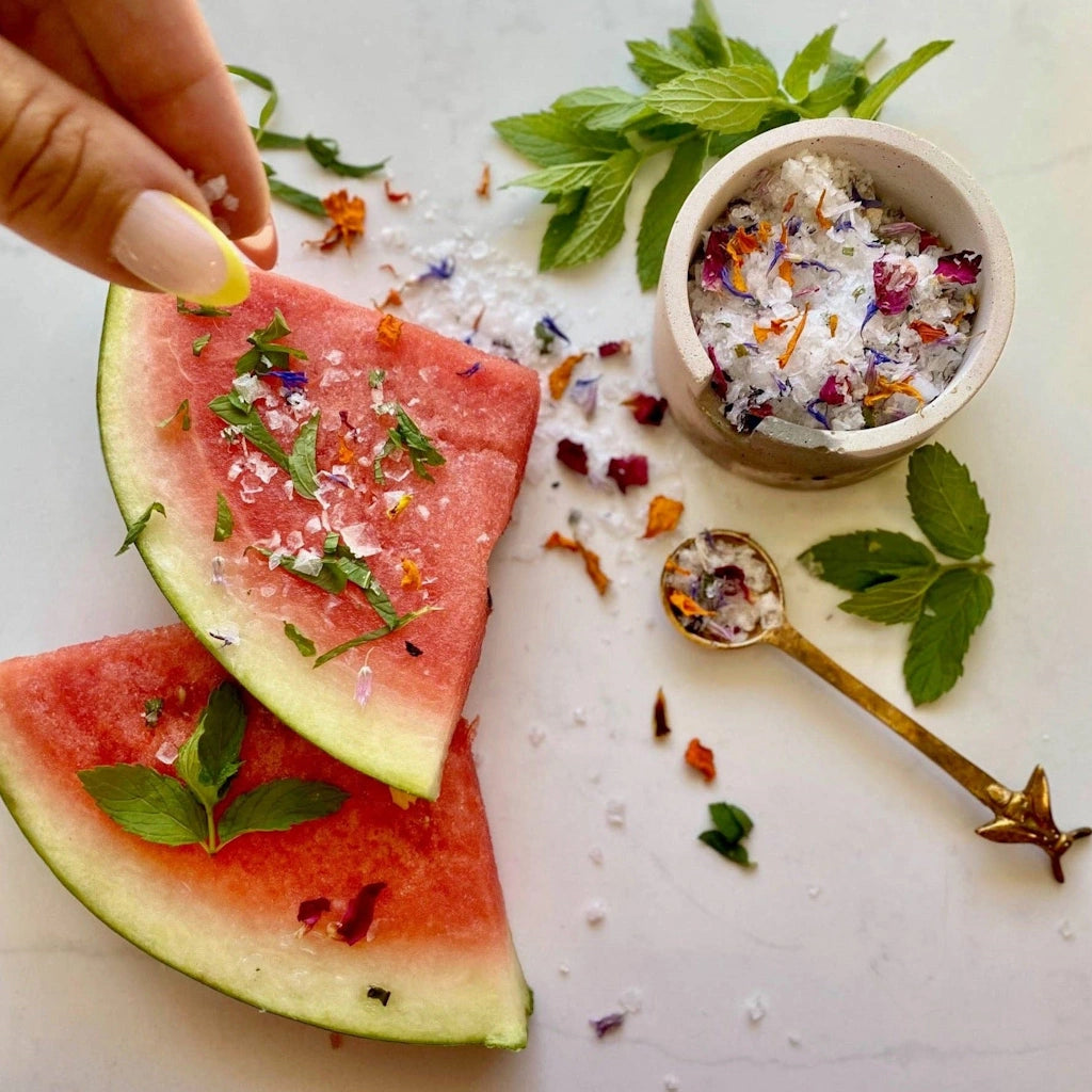 Two slices of watermelon with garnishing salt on a white surface, accompanied by a small bowl of decorative salt and a gold spoon.