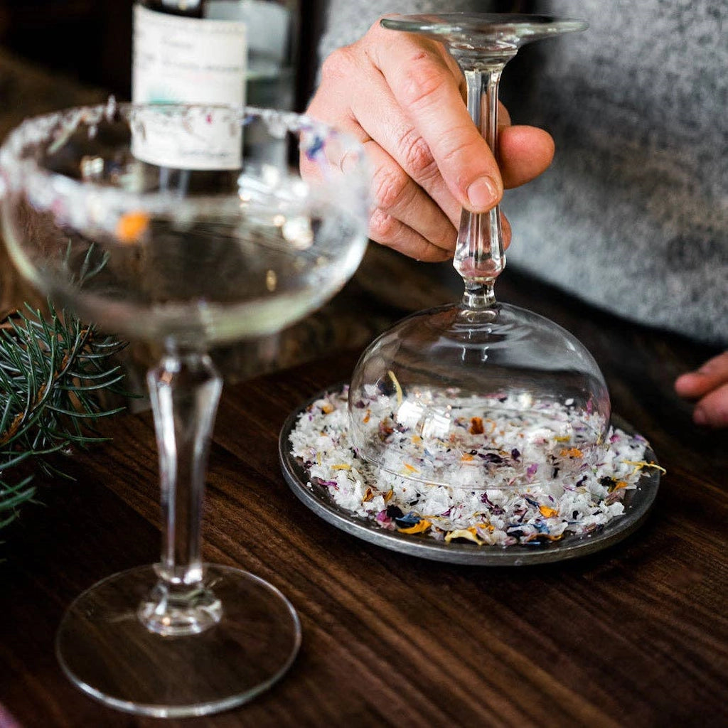 Person holding a glass over a dish with Flouwer & Co edible flower garnishing salt on a wooden table.