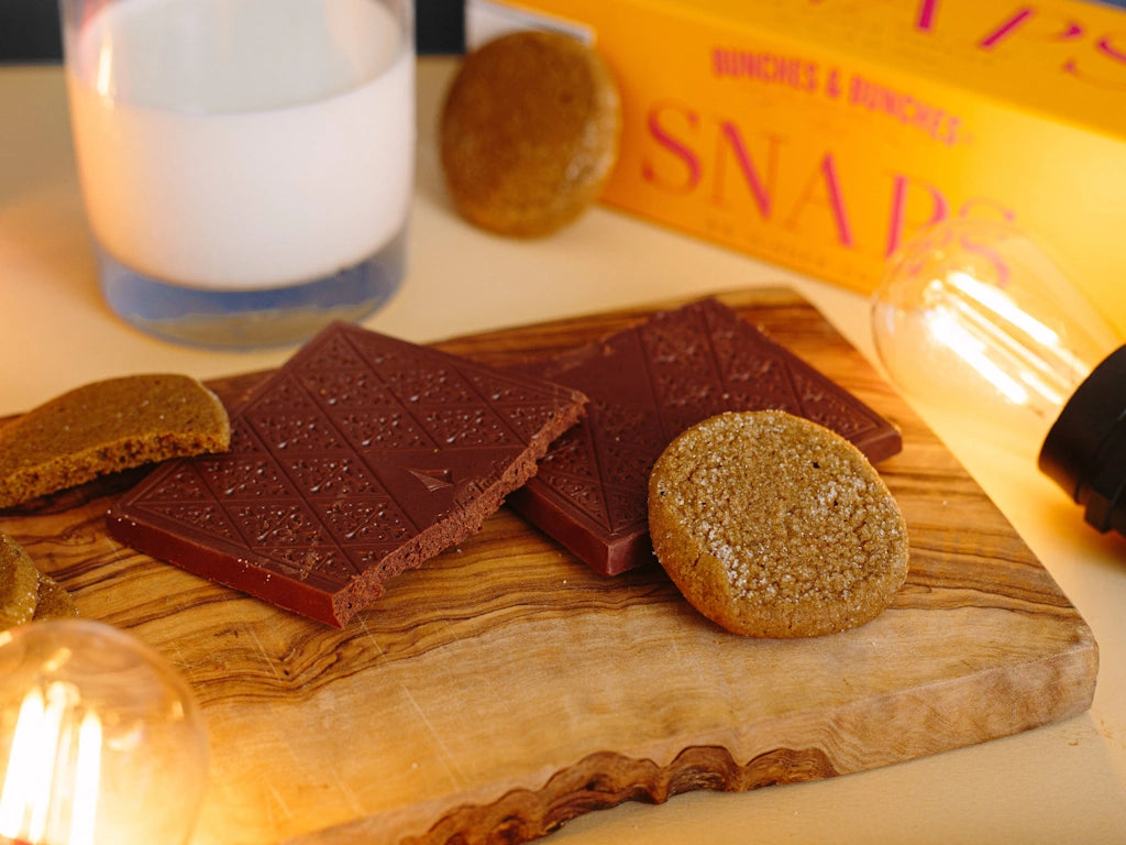 Chocolate bars and cookies on a wooden board with a glass of milk and 'Bunches & Bunchies' packaging in the background.