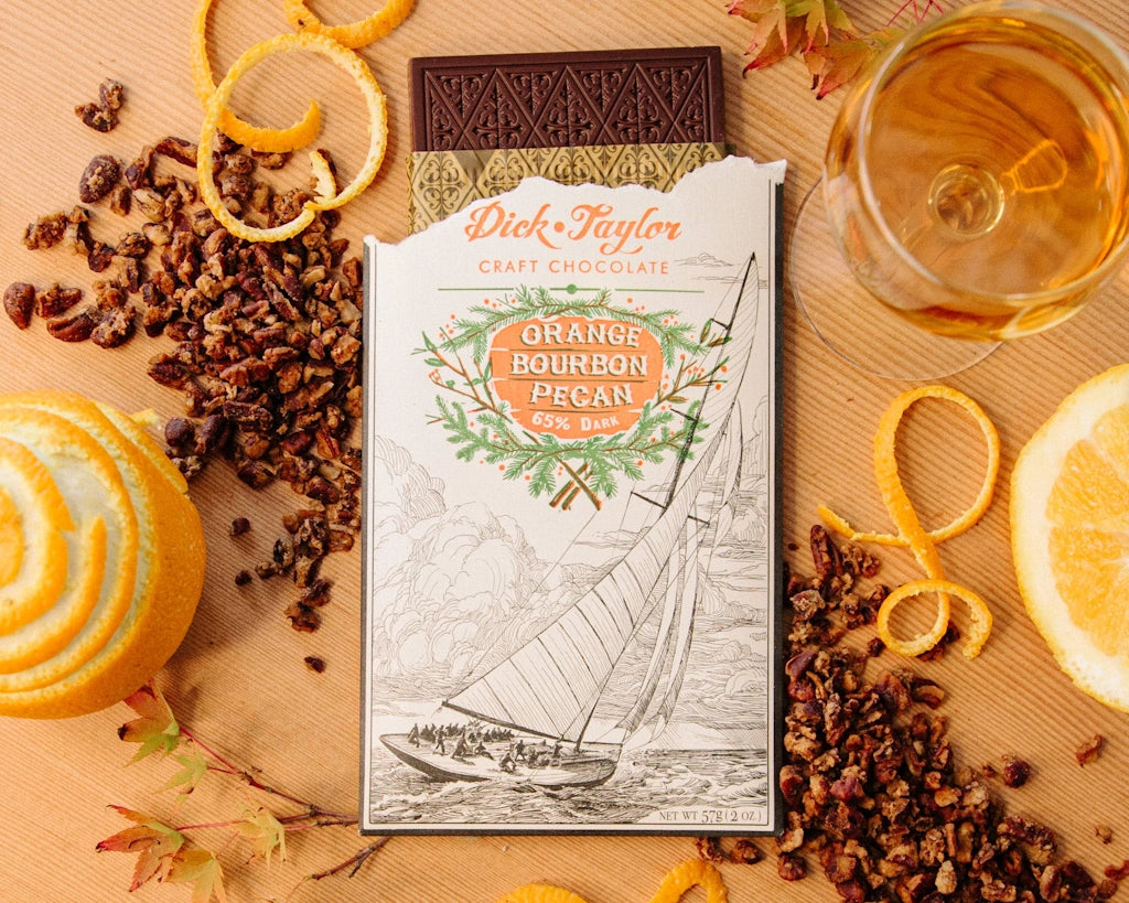 Chocolate bar with 'Orange Bourbon Pecan' flavor on a wooden surface with orange peels and chocolate pieces.