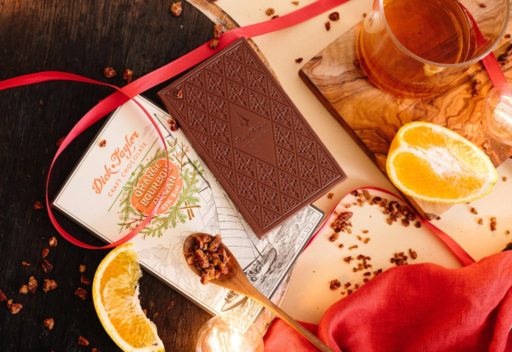 Chocolate bar with a red ribbon, orange slices, and a glass of bourbon on a wooden surface.