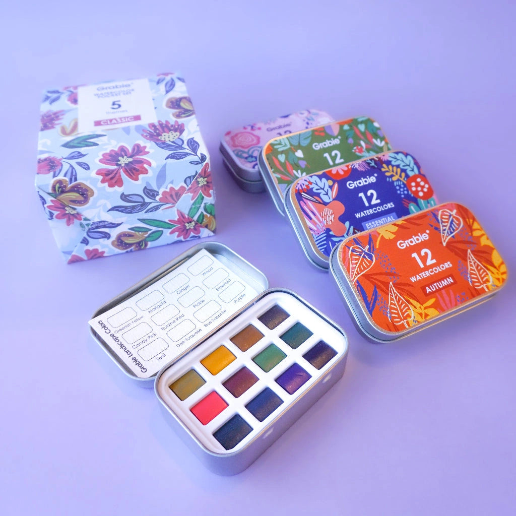 Set of watercolor pans in colorful tins on a purple background. Available from Campanula Design Studio, Seattle florist and gift basket company in the Magnolia neighborhood.