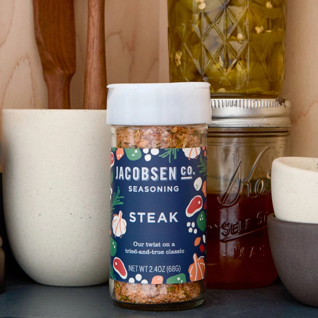 Spice jar labeled 'Jacobson Co. Steak Seasoning' on a kitchen counter with jars and a wooden spoon in the background.