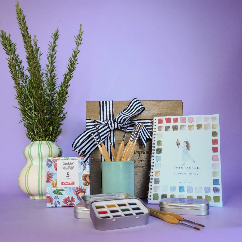 Gift box featuring art supplies including brushes, color swatches, and a plant on a purple background.  Available for sale from Campanula Design Studio, Seattle florist and gift basket company in the Magnolia neighborhood.