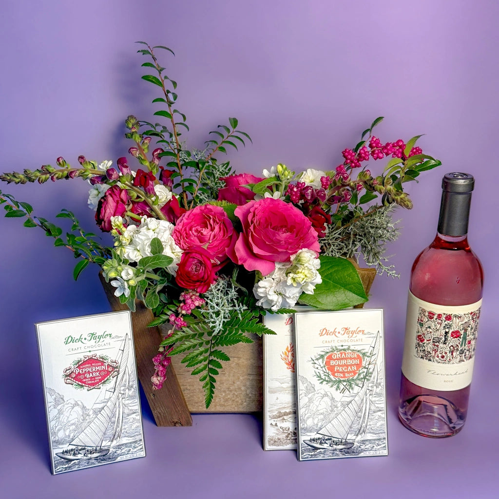 Floral arrangement in a wooden gift box with a bottle of wine and three chocolate bars on a purple background