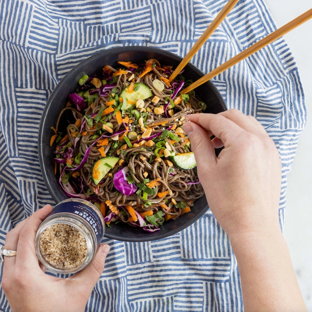 Person holding a bowl of colorful noodles and vegetables, using chopsticks and a small container Jacobsen Co Black Garlic salt.