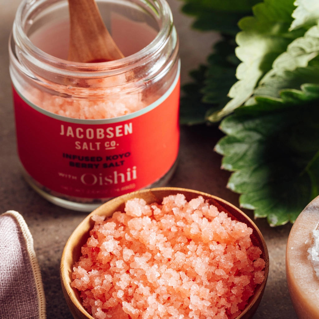 Jar of Jacobsen Salt Co. pink sea salt with a wooden spoon and a bowl of salt on a neutral background.