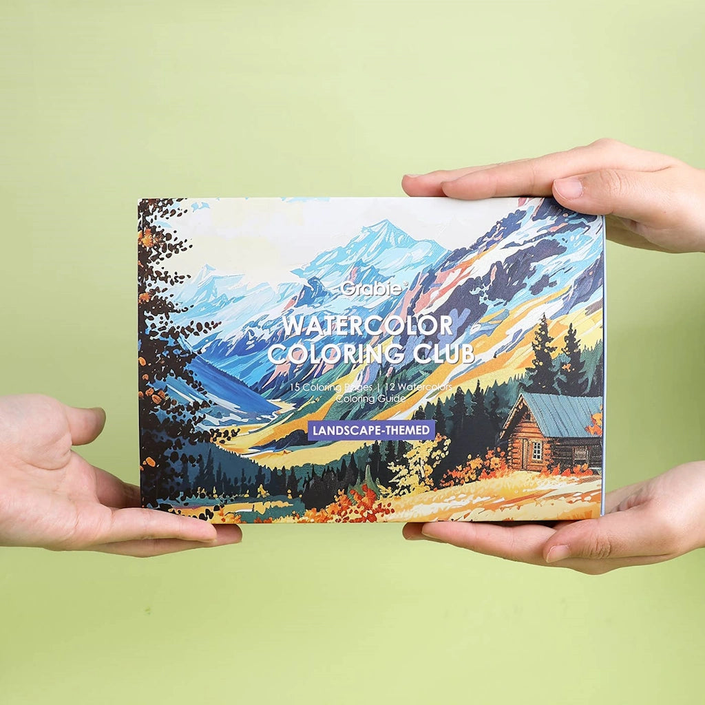 Grabie Watercolor Coloring Club landscape-themed coloring book held by two hands against a light green background