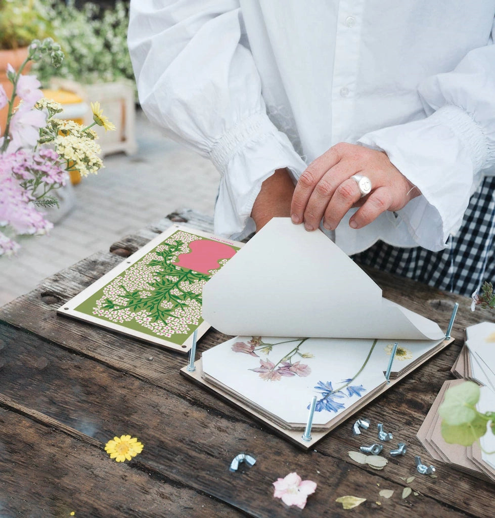 Person working with floral press and dried flowers on a wooden table. This flower press is  available for sale from Campanula Design Studio, Seattle florist and gift basket company in the Magnolia neighborhood.