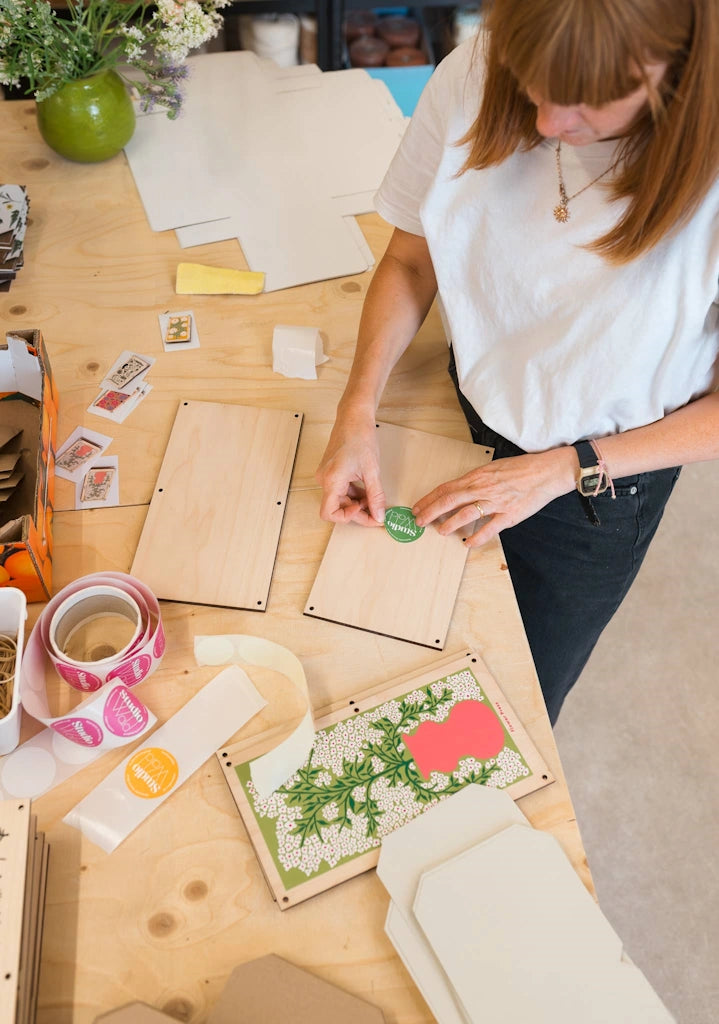 Person working on a flower press craft project with wooden boards and decorative elements on a wooden table. The flower press is available for sale from Campanula Design Studio, Seattle florist and gift basket company in the Magnolia neighborhood.