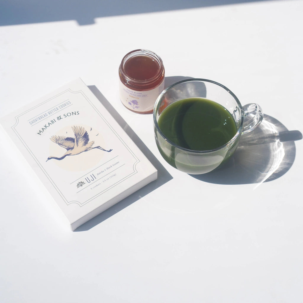 Clear glass mug with green tea, small jar, and a box of shortbread cookies on a white surface