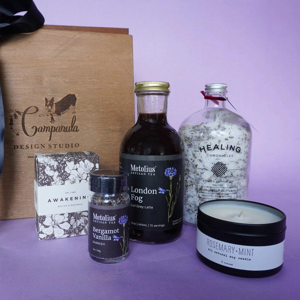 Collection of wellness products including a bottle of tea, candle, and boxes on a purple background with 'Campanula Design Studio' branding.  Available for sale from Campanula Design Studio, Seattle florist and gift basket company in the Magnolia neighborhood.