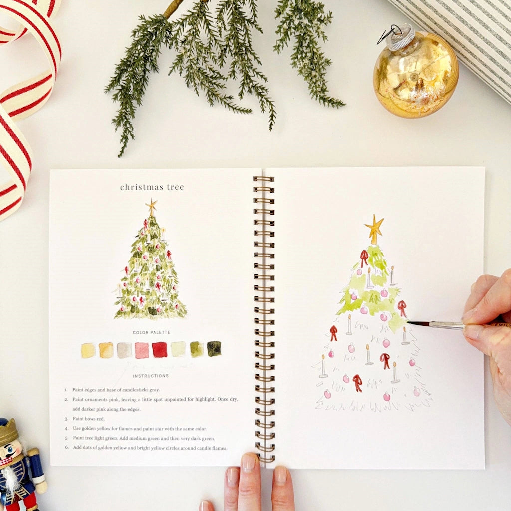 Open book with Christmas tree illustrations and a hand holding a paintbrush.