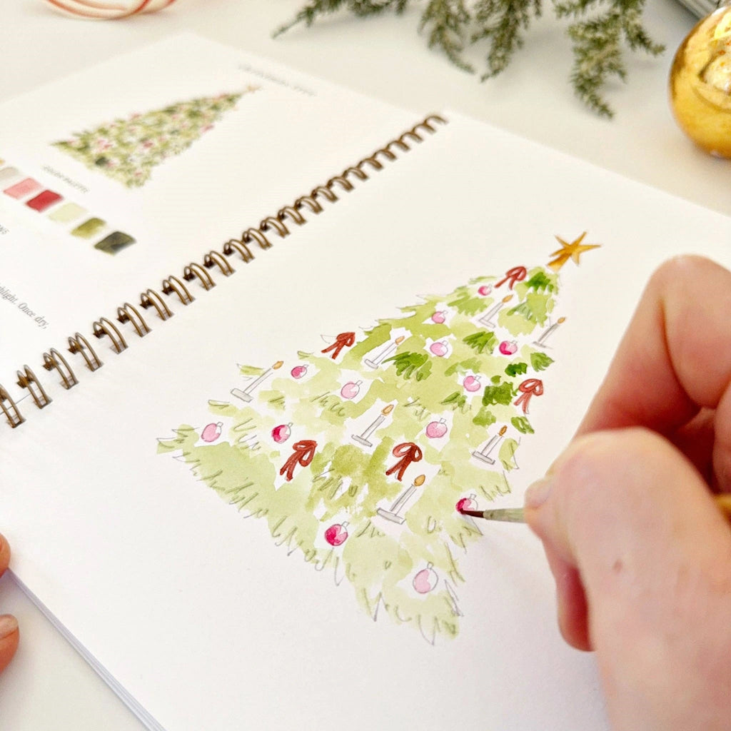 Person drawing a Christmas tree on a spiral-bound notebook with a paintbrush.