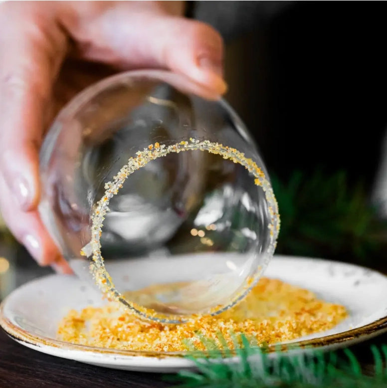 Clear glass with a decorative rim being held over a plate with orange blossom finishing sugar.