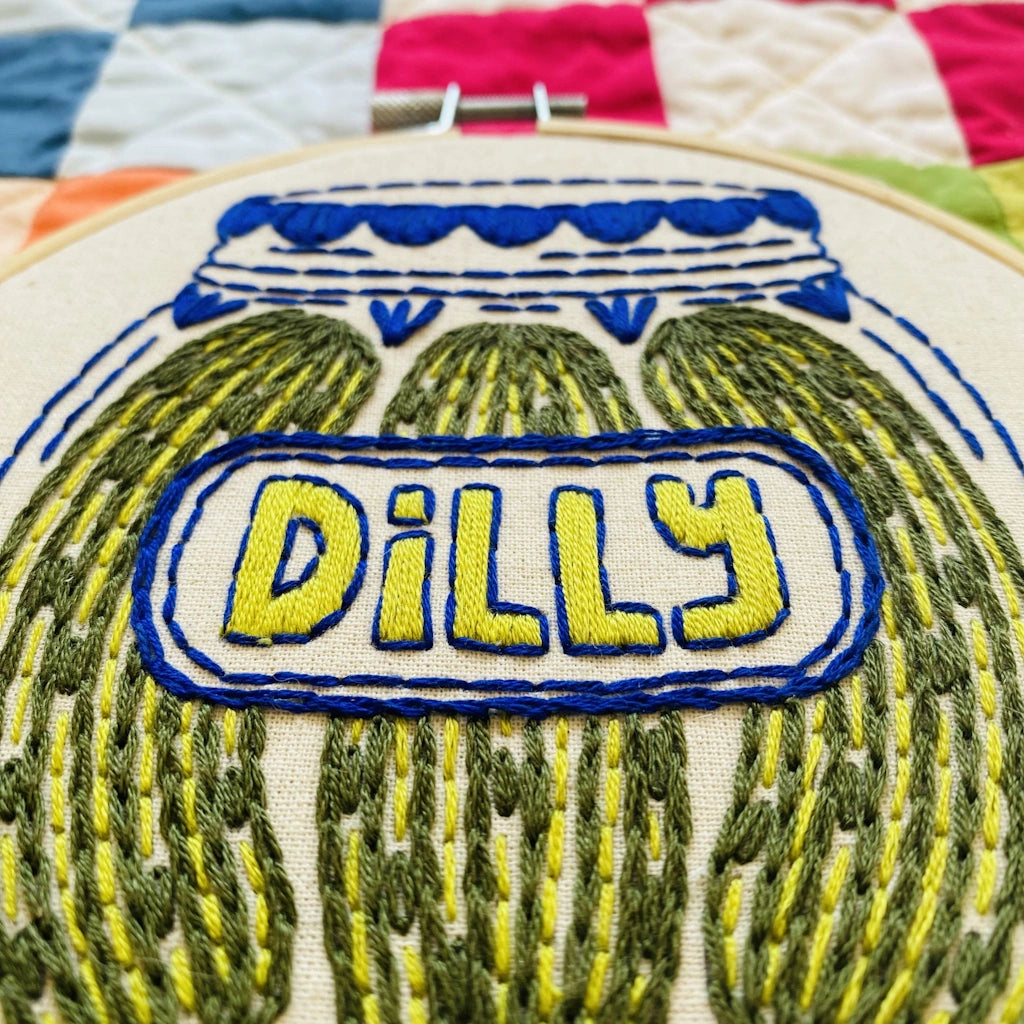 Embroidery hoop with pickles and 'Dilly' text on a patchwork background