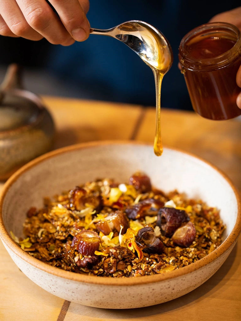 Honey being drizzled from a spoon into a bowl of granola on a wooden surface.
