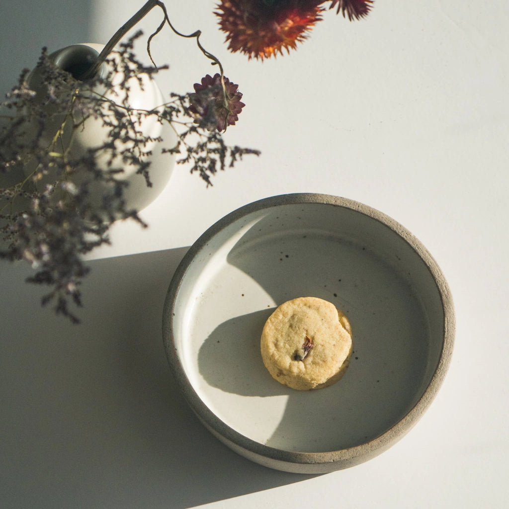 Ceramic dish with a shortbread cookie on a light surface, with a vase of flowers in the background.