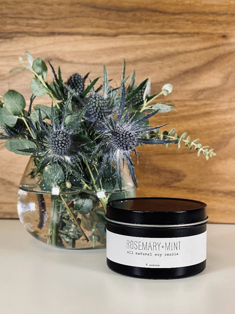 Candle named 'Rosemary + Mint' next to a glass vase with greenery on a wooden surface.