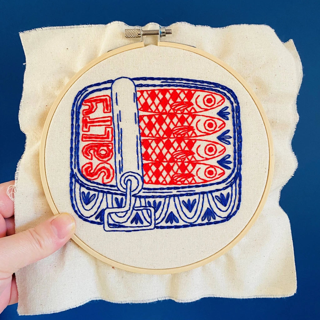 Embroidery hoop with red and blue fish pattern on beige fabric against a blue background.  Available for sale from Campanula Design Studio, Seattle florist and gift basket company in the Magnolia neighborhood.