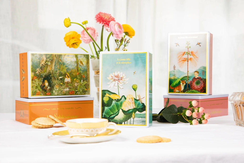 Tins of French shortbread cookies with a vase of flowers on a white background.