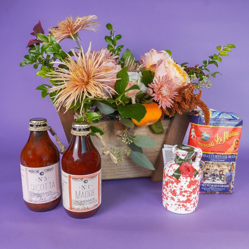 A wooden gift basket featuring a floral arrangement and Italian themed foods on a purple background.  Available for sale from Campanula Design Studio, Seattle florist and gift basket company in the Magnolia neighborhood.