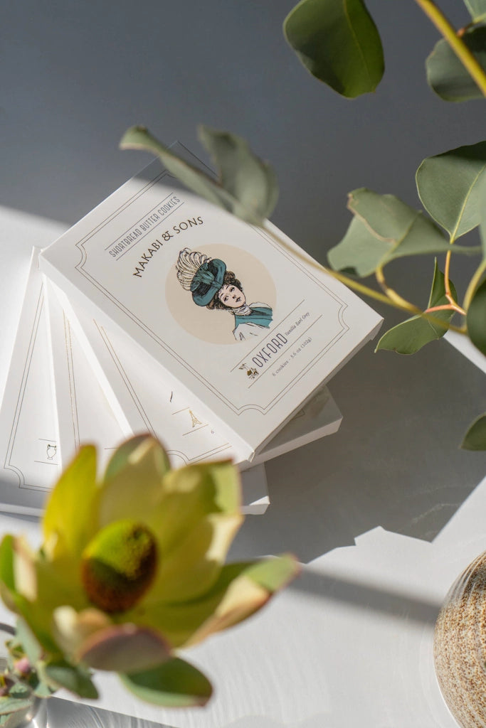 Stack of shortbread cookie boxes featuring an illustration on a gray surface with green leaves.