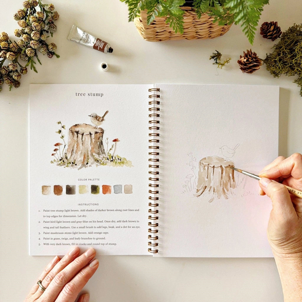 Artbook with illustrations of a tree stump and color palette, with a hand holding a paintbrush.