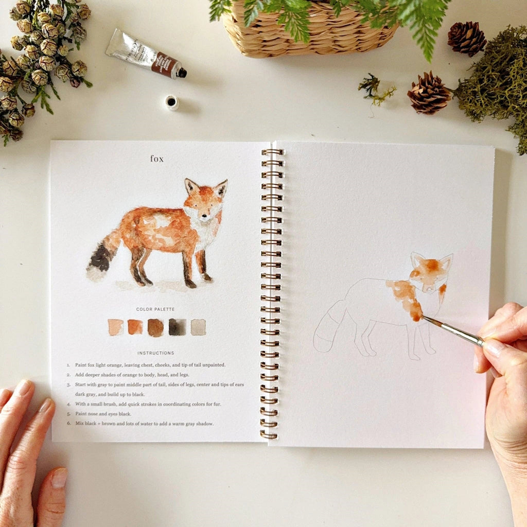 Open book with a fox illustration and color swatches, being painted by a hand.