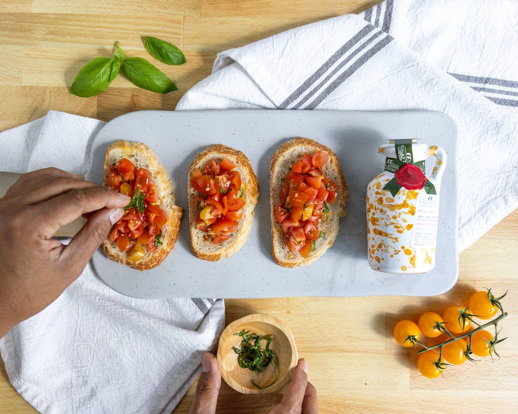 Three slices of bread with tomato salad on a marble board, with a hand reaching for a small container of olive oil.